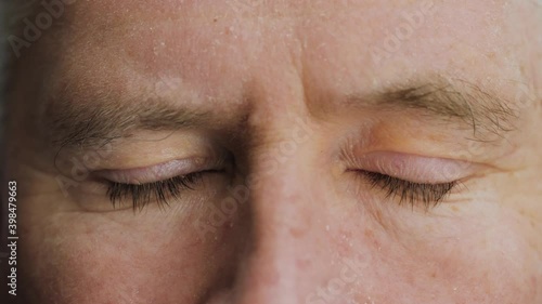 Face close up of tired elderly middle aged man closing his blue eyes. Old Caucasian mature male senior citizen with wrinkles on attractive face looking at camera. Slow motion, extreme macro, 4K.