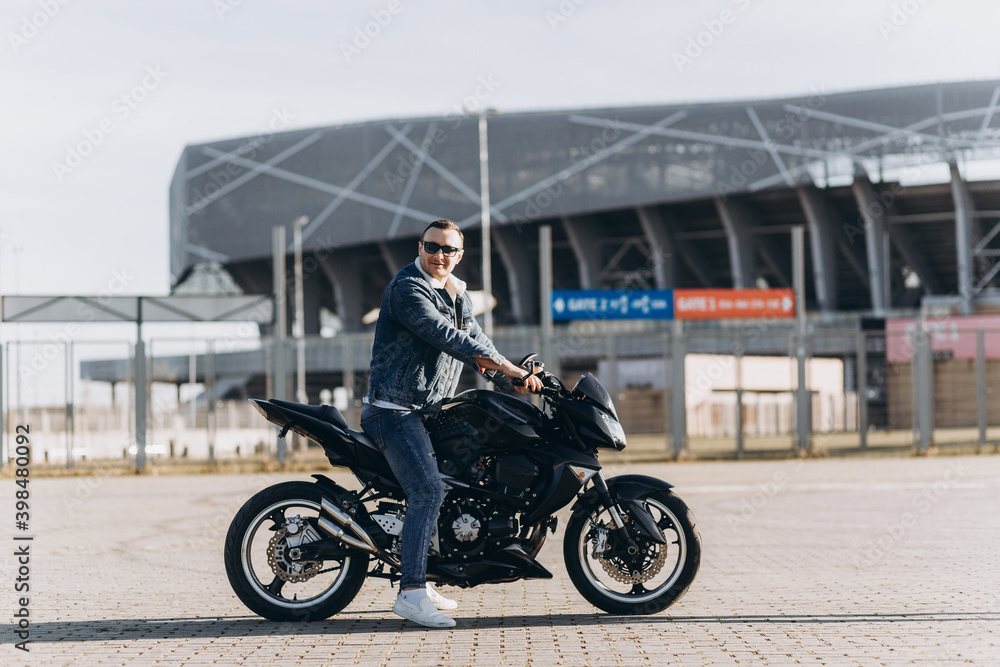 A man in denim clothes sitting on a black sports motorcycle against the backdrop of a large modern building