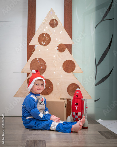 Adorable little blond kid boy  wearing Astronaut pajamas sitting near Christmas tree. Funny original creative holiday concept 