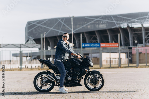 A man in denim clothes sitting on a black sports motorcycle against the backdrop of a large modern building