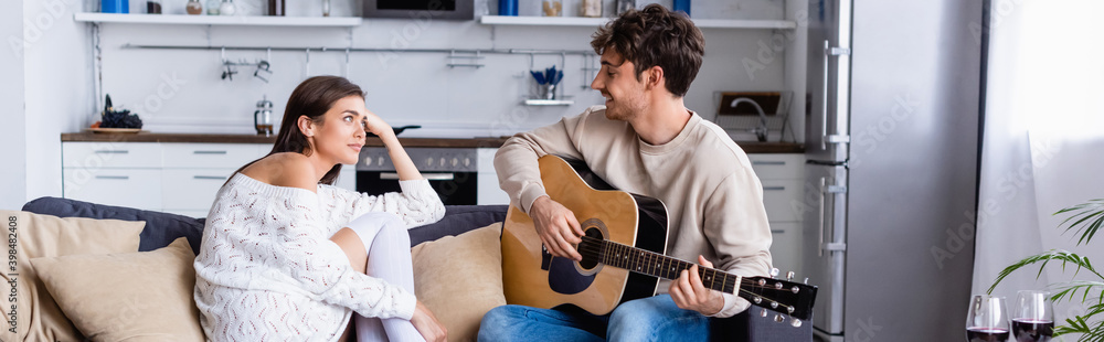 Obraz premium Smiling man playing acoustic guitar near girlfriend and glasses of wine at home, banner