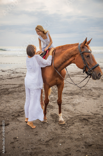 Horse riding on the beach. Cute little girl on a brown horse. Her mom standing near by. Love to animals. Mom and daughter spending time together. Outdoor activities. Mother's day.