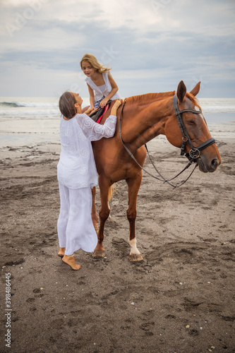 Horse riding on the beach. Cute little girl on a brown horse. Her mom standing near by. Love to animals. Mom and daughter spending time together. Outdoor activities. Mother's day.