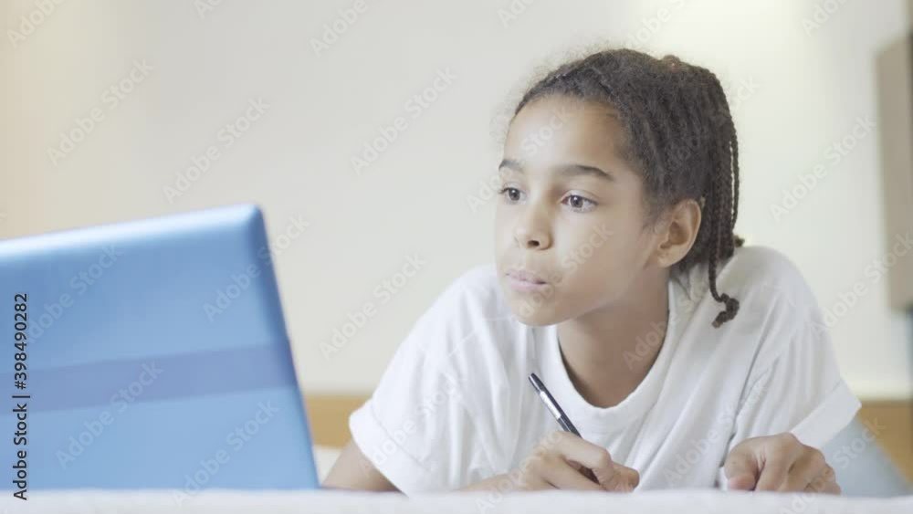 Portrait of smart concentrated schoolgirl doing homework lying on bed with laptop. Portrait of cute African American teenager studying online at home. Education and intelligence concept. Portrait of smart concentrated schoolgirl doing homework lying on bed with laptop. Portrait of cute African American teenager studying online at home. Education and intelligence concept.