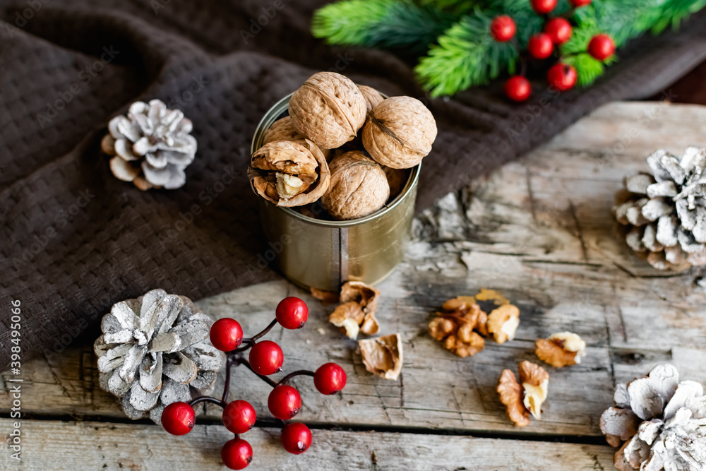 Fototapeta premium Walnuts in a tin, whole and split, next to the filling and shell. Presented in a New Year's composition with cones, red berries and a sprig of a Christmas tree. Home storage of winter preparations