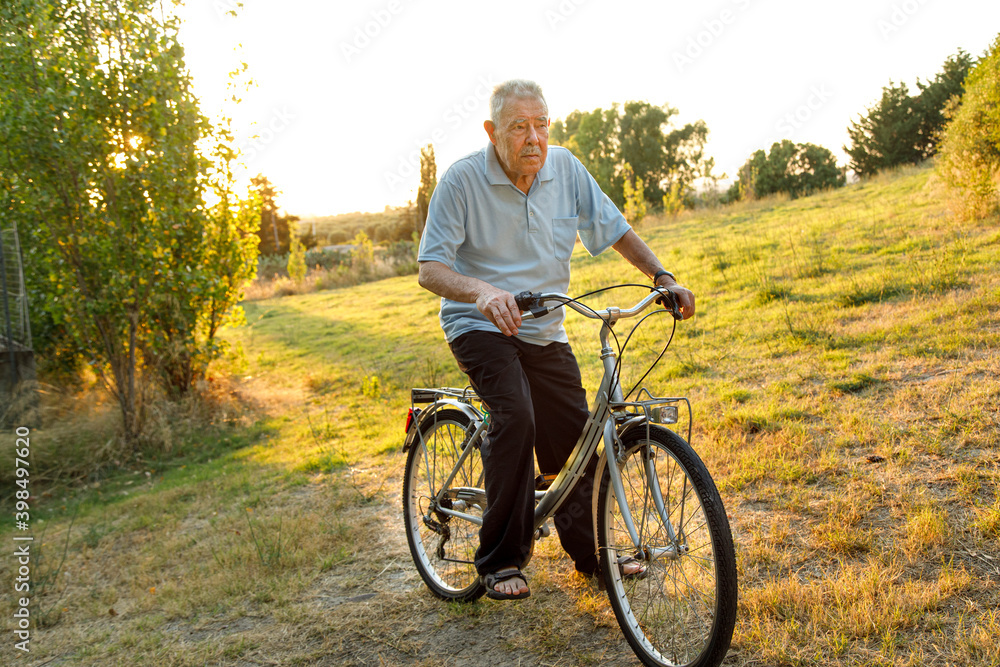 Naklejka premium Signore ottantenne sta in sella alla sua bicicletta in un prato verde al tramonto