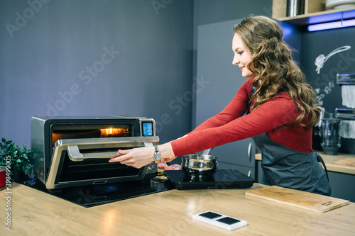 A beautiful woman opens the door of a mini oven to check the readiness of the dish.