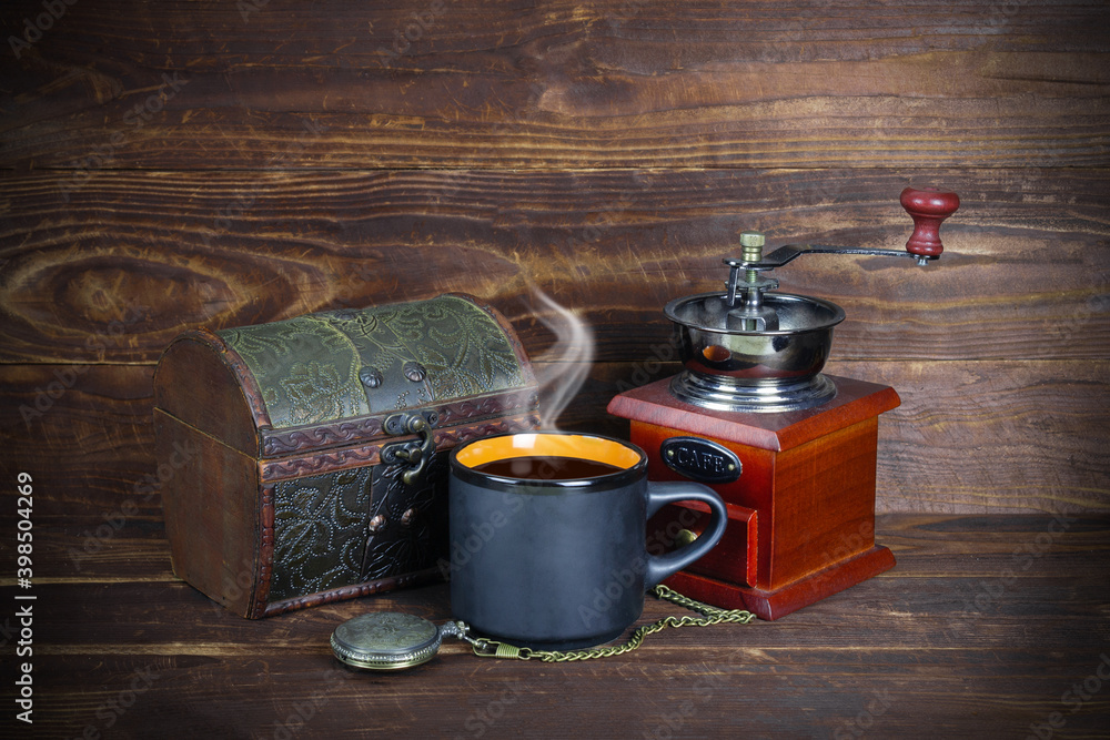 Retro jewelry box, black mug with coffee and steam above it, pocket watch with chain, vintage coffee grinder with handle on brown wooden plank background.