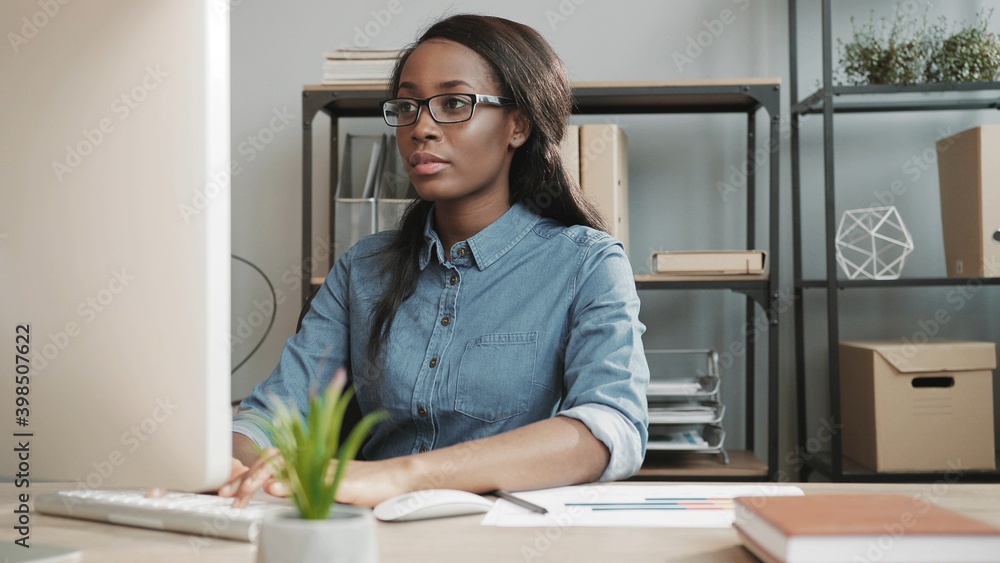 Young beautiful african american girl in glasses and denim shirt sitting in modern office working at computer making notes in paper documents. Young beautiful african american girl in glasses and denim shirt sitting in modern office working at computer making notes in paper documents.