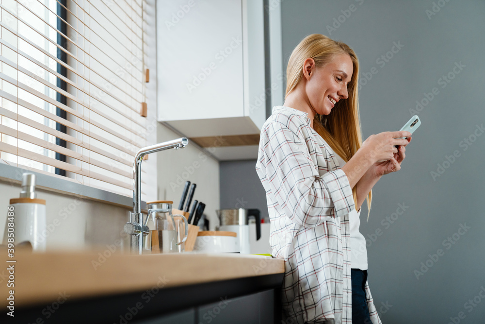 Blonde happy woman using mobile phone and smiling at home kitchen Blonde happy woman using mobile phone and smiling at home kitchen