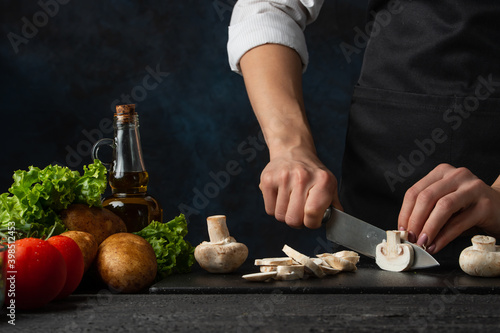 Macro shot view of chef in black apron cuts with knife mushrooms on the chopped board for cooking soup on dark blue background. Backstage of preparing meal. Healthy dish. Food concept. Frozen motion.