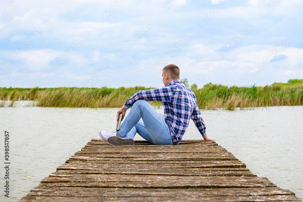 Man in blue shirt sitting on wooden dock and looking in lake. He is holding book in left hand.