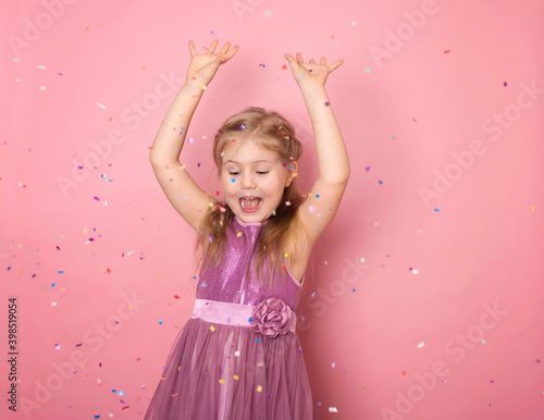 Happy girl celebrating on a pink background. Happy child with hans up looking at confetti rain. People, holidays, emotion and glamour concept.