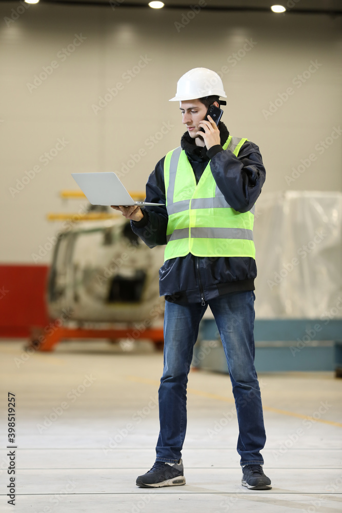 Young man, factory engineer in a work dress and white hard hat holding smartphone and laptop organizing and controlling work process in a industrial factory.