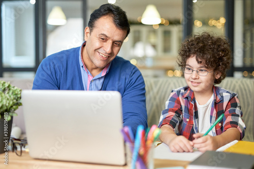 Partnership. Portrait of cheerful middle aged hispanic father spending time with his son. Little boy sitting at the desk together with his dad and doing homework, using laptop
