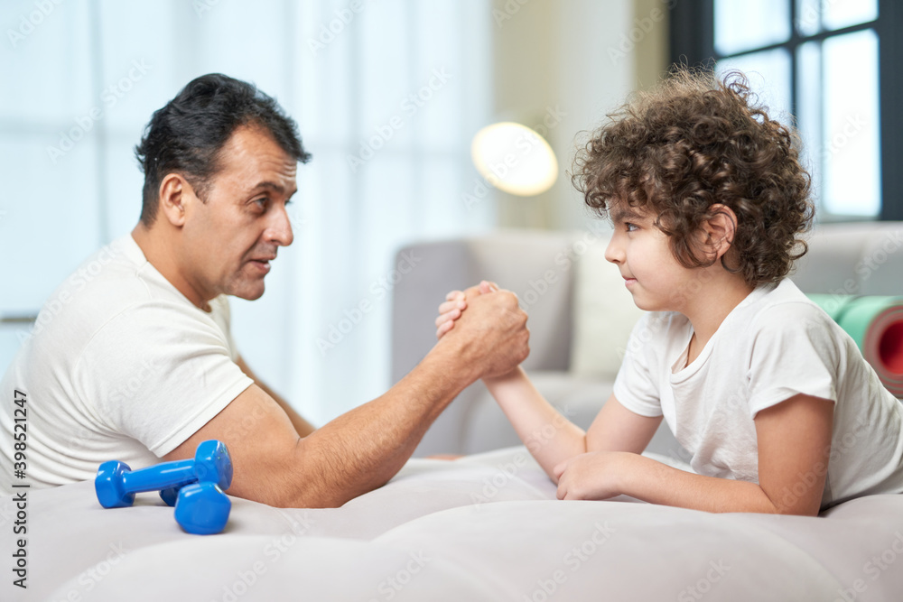 Determination. Cute latin boy and his father having fun together at home while armwrestling lying on the couch Determination. Cute latin boy and his father having fun together at home while armwrestling lying on the couch