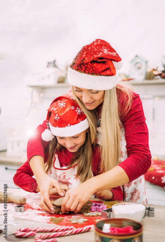 A young mother and her teenage daughter are standing in the kitchen and  together preparing dough for baking, laughing and smiling in red sweaters and hats. Vertical photo