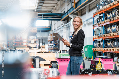 Happy female entrepreneur holding digital tablet looking away while standing in factory
