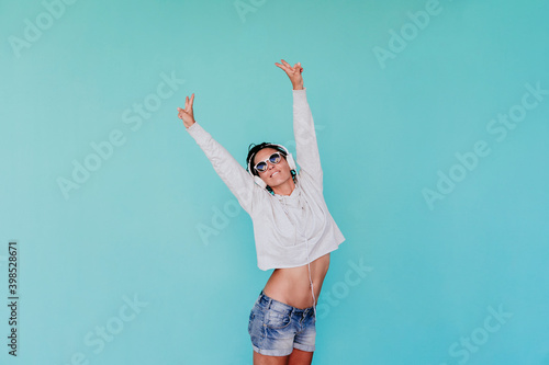 Happy fashionable woman listening music while standing with arms raised against turquoise background photo