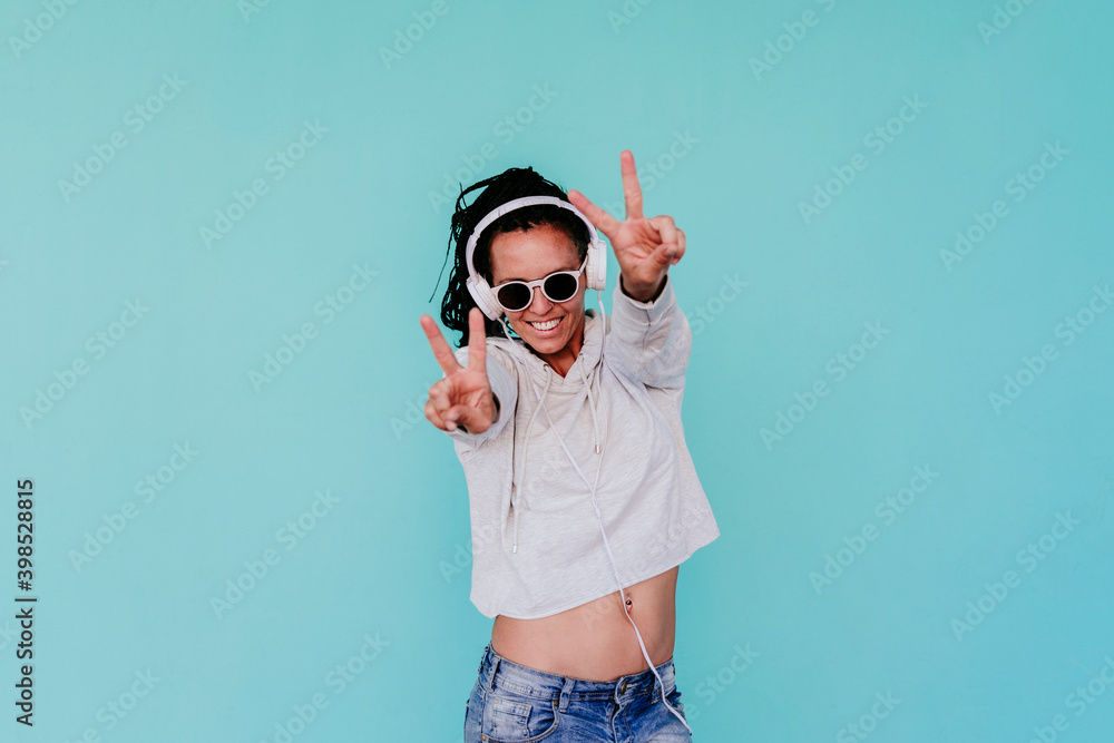 Happy fashionable woman showing peace sign while listening music through headphones against turquoise background