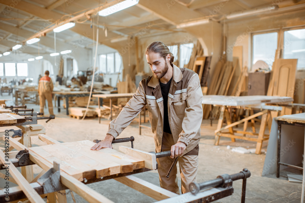 Handsome carpenter in uniform gluing wooden bars with hand pressures at the carpentry manufacturing Handsome carpenter in uniform gluing wooden bars with hand pressures at the carpentry manufacturing