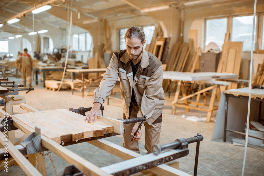 Handsome carpenter in uniform gluing wooden bars with hand pressures at the carpentry manufacturing Handsome carpenter in uniform gluing wooden bars with hand pressures at the carpentry manufacturing