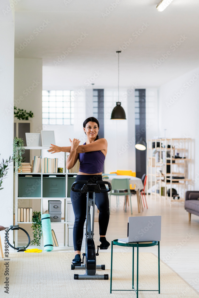 Young female athlete doing stretching exercise while sitting on exercise bike at home Young female athlete doing stretching exercise while sitting on exercise bike at home