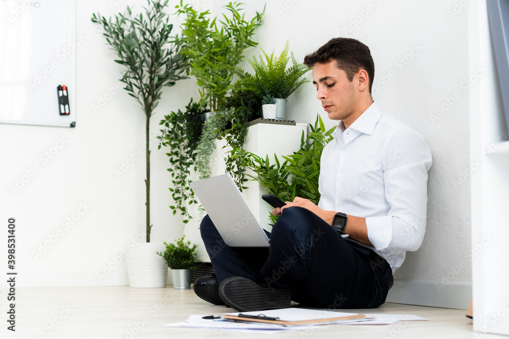 Male professional using phone while sitting with laptop against white wall at office Male professional using phone while sitting with laptop against white wall at office