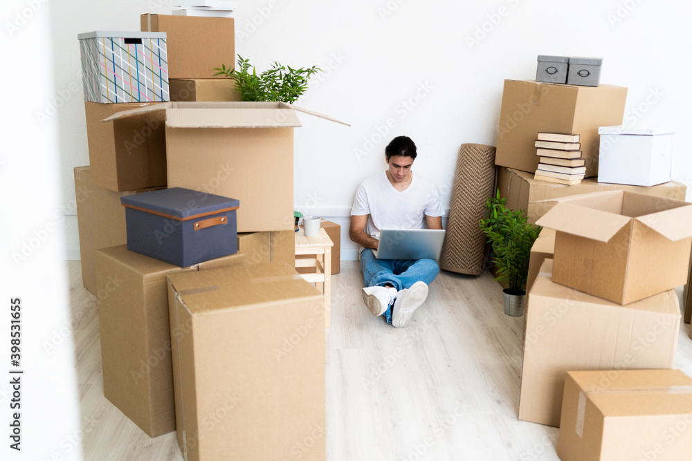 Man working on laptop surrounded by boxes while sitting in new apartment Man working on laptop surrounded by boxes while sitting in new apartment
