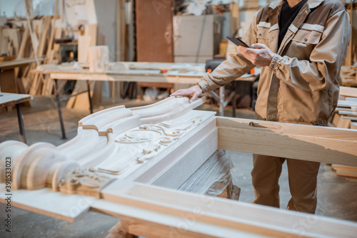 Handsome carpenter working on wooden carvings using a smart phone at the carpentry manufacturing. Concept of using mobile technologies in carpentry