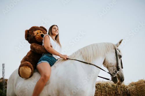 Beautiful young woman riding horse with large teddy bear behind photo