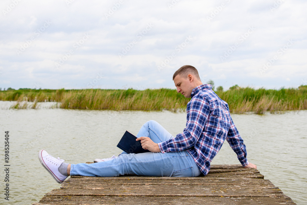Man in blue shirt reading book on wooden dock on lake.