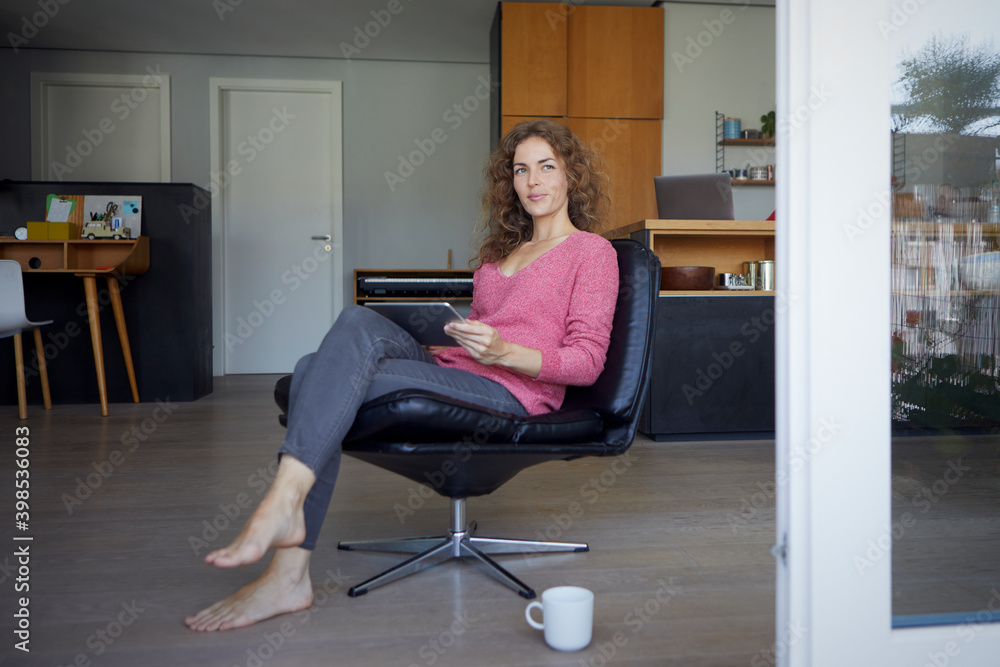 Woman looking away while using digital tablet sitting on chair at home Woman looking away while using digital tablet sitting on chair at home