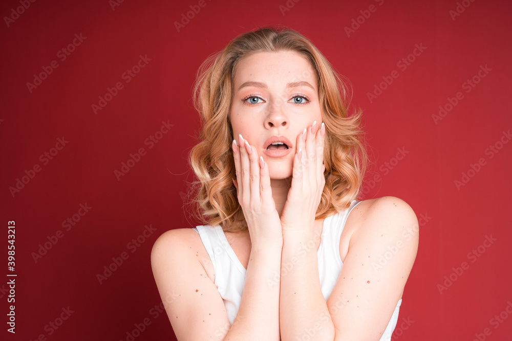Blond woman with curly hair holds her head with hands in white tank top isolated on red background. Blond woman with curly hair holds her head with hands in white tank top isolated on red background.