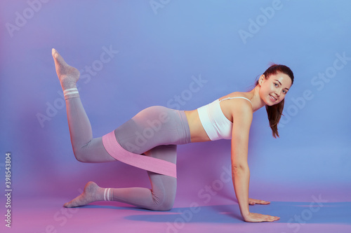 Smiling sporty Caucasian woman has exercises with rubber resistance band, trains buttocks, raising feet, works on muscles, dressed in top and leggings, poses indoor against neon background.