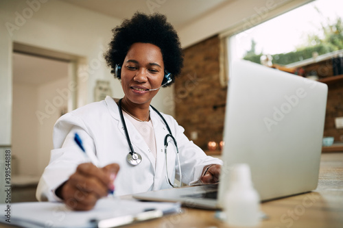 Happy African American doctor taking notes while working on laptop at her office.