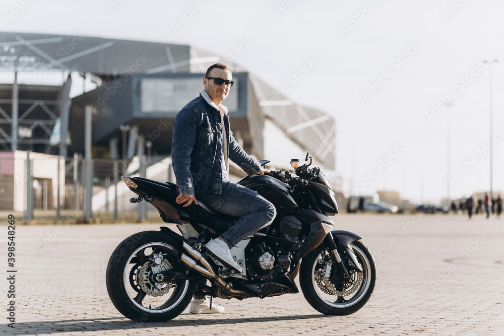 A man in denim clothes sitting on a black sports motorcycle against the backdrop of a large modern building