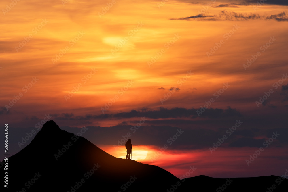 Beautiful dark golden sunset. Silhouette of a tourist on a hill stands and looks at the huge sun.