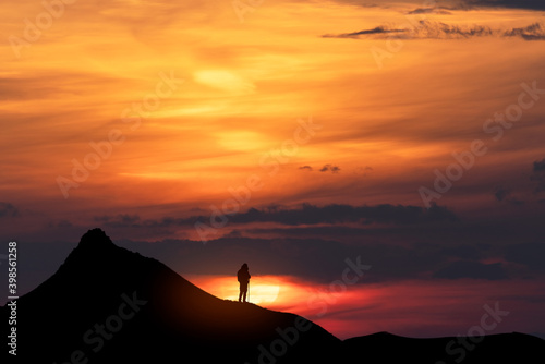 Beautiful dark golden sunset. Silhouette of a tourist on a hill stands and looks at the huge sun.