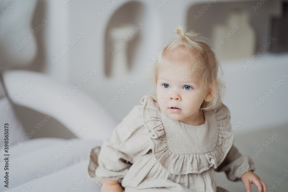 beautiful little girl of European appearance blonde with a ponytail. In a beige dress posing stands playing near the sofa and looks away. Bright and cozy apartment in the city center. beautiful little girl of European appearance blonde with a ponytail. In a beige dress posing stands playing near the sofa and looks away. Bright and cozy apartment in the city center.