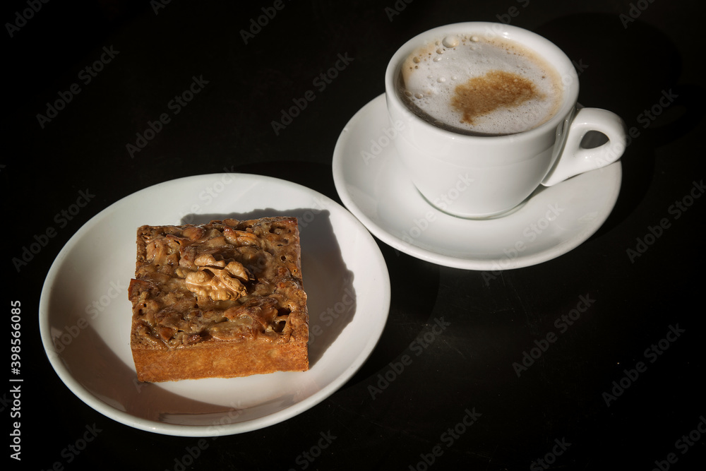 walnut cake on a white plate next to a Cup of cappuccino, Sunny photo. walnut cake on a white plate next to a Cup of cappuccino, Sunny photo.