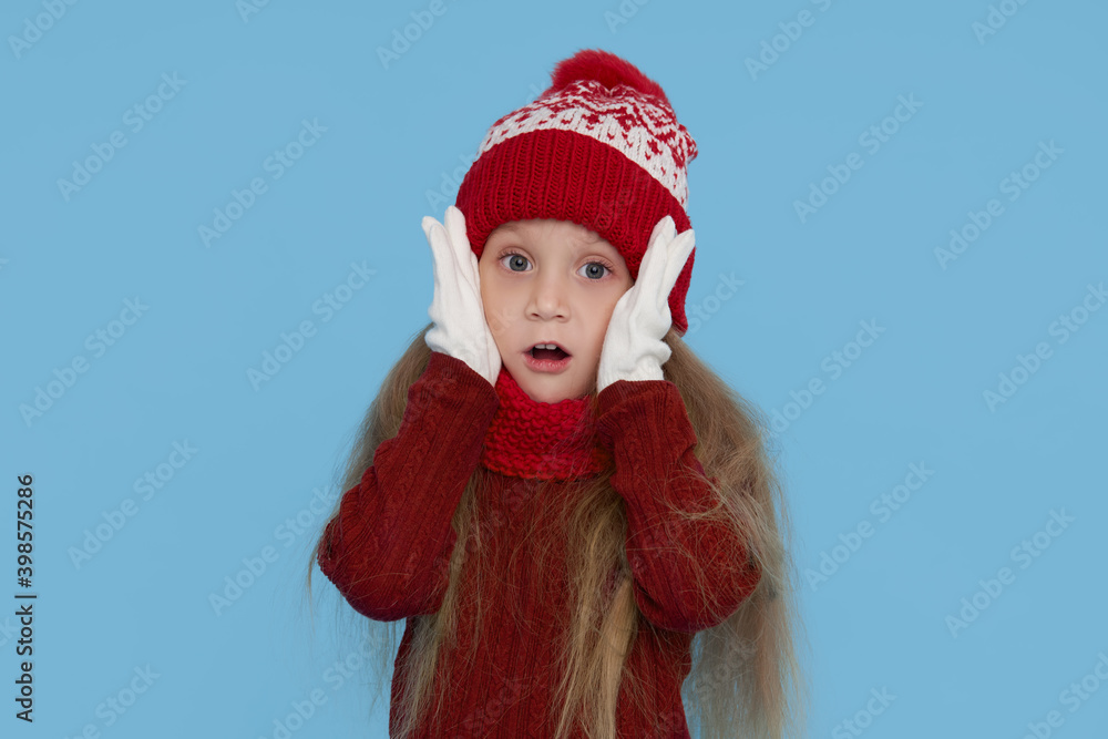 Winter portrait of happy child girl wearing knitted red hat, snood and sweater. Wow face. Blue background. childrens winter entertainment