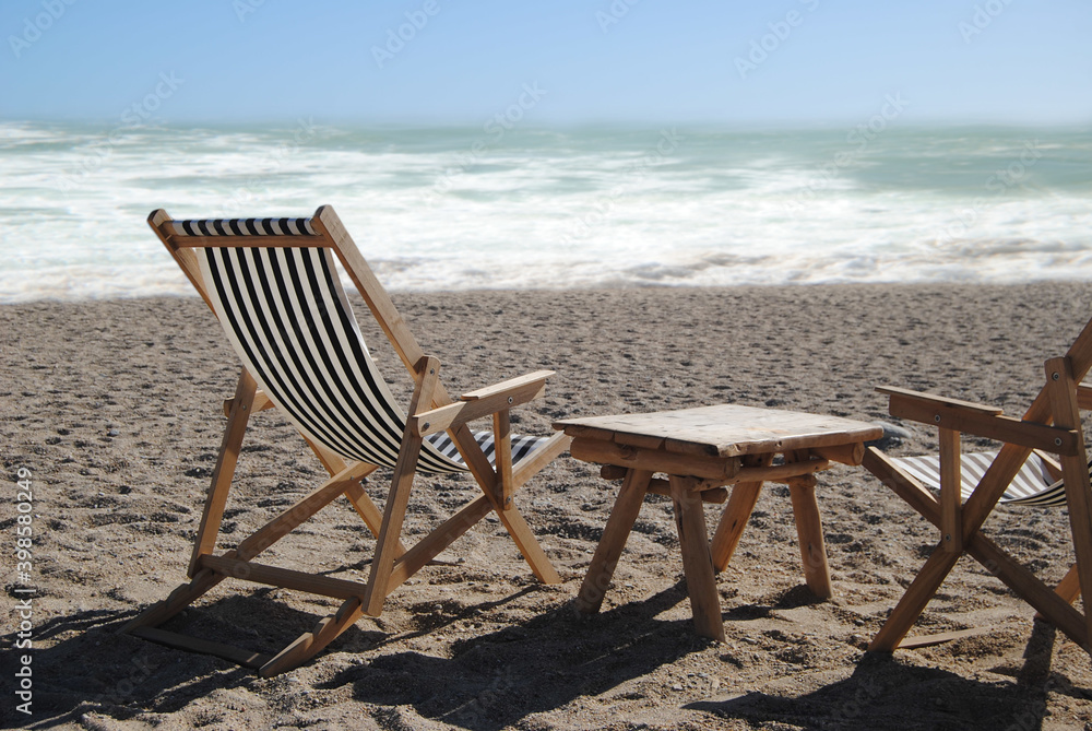 Duas espreguiçadeiras de praia e uma mesa, colocadas  no meio de uma praia com o oceano de fundo em segundo plano desfocado, espreguiçadeiras com tecido ás riscas brancas e preto