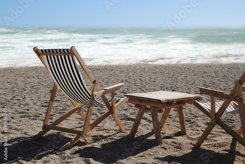 Duas espreguiçadeiras de praia e uma mesa, colocadas  no meio de uma praia com o oceano de fundo em segundo plano desfocado, espreguiçadeiras com tecido ás riscas brancas e preto © ajcsm