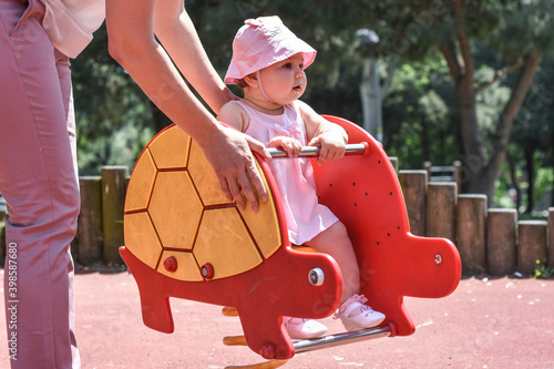Mother swinging her cute adorable baby daughter in rocking turtle in park (garden) Happy family moments concept. Family time. Outdoor activity at a playground in a summer days. Happy childhood. 