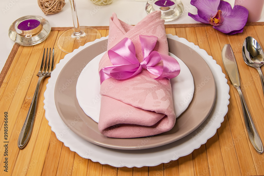 Table setting. Beautiful dishes and a pink napkin with a satin ribbon on a wooden stand with a crystal glass, candles and flowers. White background. Table setting. Beautiful dishes and a pink napkin with a satin ribbon on a wooden stand with a crystal glass, candles and flowers. White background.