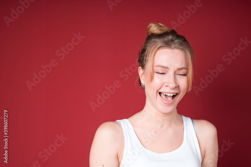 Russian blond woman with bun and white tank top laughing isolated on red background.