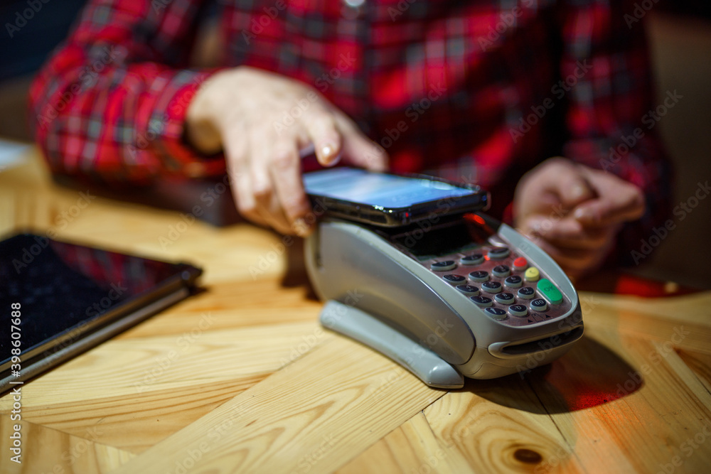 Fototapeta premium Contactless payment. Nfs payment by phone. Client of cafe or restaurant holding smartphone over electronic payment machine while using mobile app to pay. Non cash payment. NFC technology