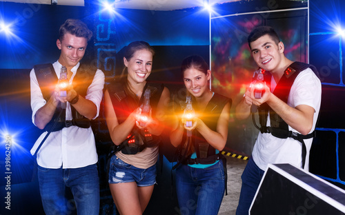 Four cheerful young male an female posing with laser pistols in their hands in dark laser tag room