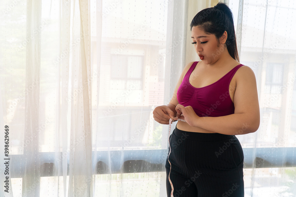 Obesity unhealthy concept. Overweight woman wear sport bar with measure tape on body pinches fat on her belly large size while standing beside windows feeling unhappy. Obesity unhealthy concept. Overweight woman wear sport bar with measure tape on body pinches fat on her belly large size while standing beside windows feeling unhappy.